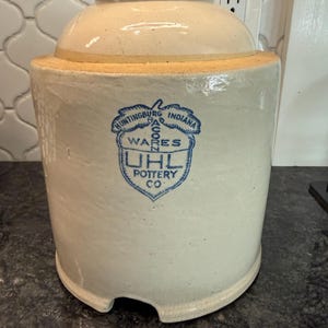 May include: Off-white stoneware crock with a matching lid. The crock features a blue emblem with the text "Huntingburg Indiana" and "UHL Pottery Co." The crock has a rounded shape with a handle-less lid and a small notch at the base.