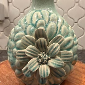 May include: A decorative ceramic vase in shades of teal and white. The vase features a textured surface with a large, three-dimensional flower design on the front. The flower has detailed petals and a textured center, adding visual interest.