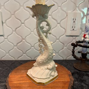 May include: A cream-colored ceramic candlestick holder with a dolphin-shaped stem and gold trim. The candlestick holder is on a round wooden base. The top of the candlestick holder has a gold rim.