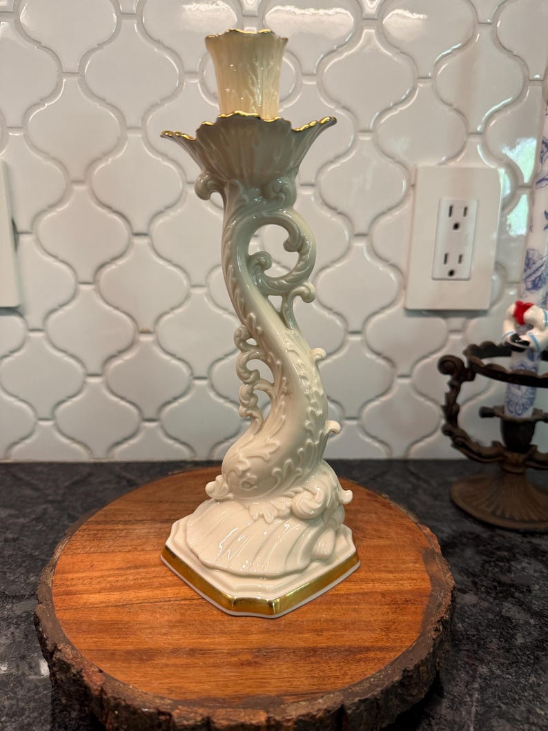 May include: A cream-colored ceramic candlestick holder with a dolphin-shaped stem and gold trim. The candlestick holder is on a round wooden base. The top of the candlestick holder has a gold rim.
