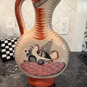 May include: A hand-painted ceramic pitcher with a bird design. The pitcher features a red handle and rim, with a cream-colored body adorned with a bird and floral motif. Gray and red accents decorate the neck. The base is red.
