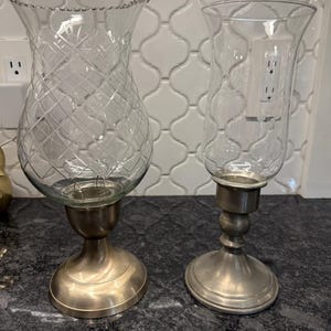 May include: Two clear glass candle holders with metal bases. One has a textured diamond pattern on the glass and a gold-toned base. The other has a smooth glass and a silver-toned base. Both are designed to hold candles.