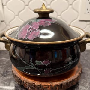 May include: A black enamel cooking pot with a lid, featuring a floral design in shades of purple and green. The pot has gold-colored handles and trim. The pot is sitting on a wooden coaster.