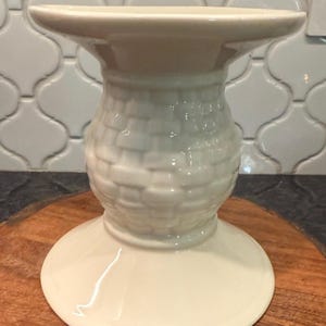 May include: Off-white ceramic candle holder with a basket weave design on the central column. The holder has a wide base and a flared top, suitable for holding a candle. The piece is set on a wooden surface.