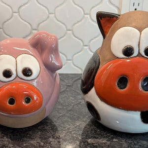 May include: Two ceramic animal figurines: a pink pig and a cow. The pig has a large orange snout, white eyes, and black pupils. The cow has a brown head, black and white spots, and a large orange snout. Both are cartoonish.