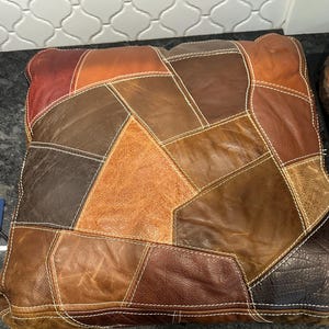May include: A patchwork leather pillow with brown, tan, and red leather pieces sewn together. The pillow has white stitching around each piece of leather.