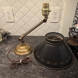 May include: An antique brass desk lamp with a black lampshade. The lamp has an adjustable arm and a round base. The black lampshade has a gold leaf design around the bottom edge. The lamp is unplugged and sitting on a dark countertop.