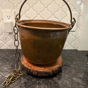 May include: A round, metallic copper-colored pot with a smooth, curved body and a wide opening. It features a sturdy, arched metal handle attached to the sides and a long, dangling metal chain. The pot rests on a circular, natural wood slice base.