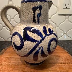 May include: A ceramic pitcher with a beige speckled body and a dark blue, stylized floral design. The pitcher has a curved handle and a spout. The rim and handle are also dark blue.