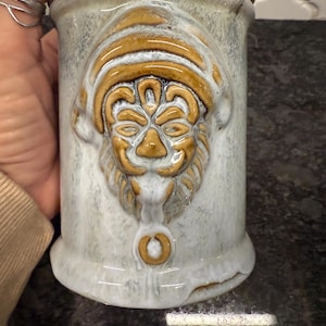 May include: A ceramic stein with a detailed relief of a face, featuring a tan and white color scheme. The stein has a lid and a handle, and the face is adorned with a hat. The stein is approximately 6 inches tall.