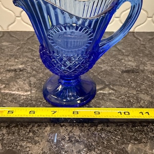 May include: A cobalt blue glass creamer with a handle and a decorative embossed design. The creamer features a diamond pattern on the base and a detailed emblem. The creamer is approximately 4 inches tall.