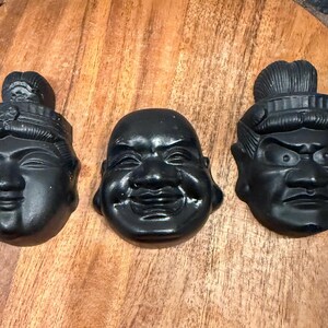 May include: Three black decorative face sculptures with distinct expressions. The faces are arranged on a wooden surface. The sculptures include a smiling Buddha-like face, a stern face, and a serene face with a headdress.