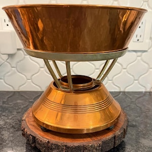 May include: A copper fondue pot with a wide bowl and a decorative base. The pot sits on a wooden base with a natural edge. The base has a copper-colored, conical-shaped body with horizontal ridges. The pot is supported by a metal frame.