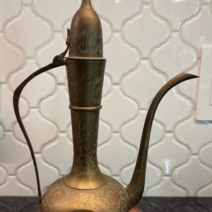 May include: A tall, antique-style brass ewer featuring an elongated neck, a curved handle, and a long, slender spout. The metal surface is decorated with intricate, hand-etched floral and leaf patterns throughout the body and the lid.