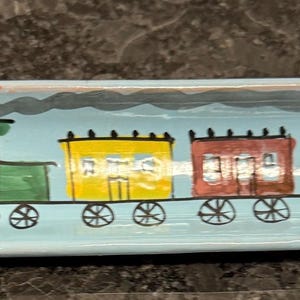 May include: A hand-painted, rectangular ceramic dish with a light blue background. The design features a green train engine pulling four colorful train cars: yellow, red, orange, and green. Black smoke billows from the engine's smokestack.