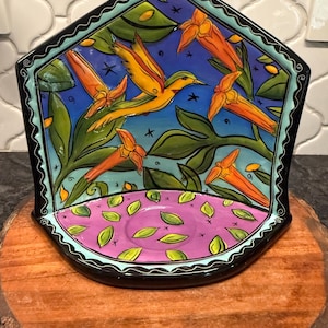 May include: A decorative ceramic tray with a hummingbird and floral design. The tray features a black border, a blue background, and a pink base. The hummingbird is yellow and orange, and the flowers are orange.