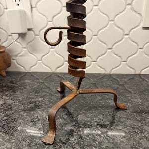 May include: A rustic, wrought iron candle holder with a spiral design. The candle holder has a three-legged base and decorative curled accents. The metal has a weathered, brown finish, adding to its vintage appeal. A unique home decor item.