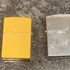 May include: Four vintage Zippo lighters in a row. The first is a dark gray, textured lighter with the Zippo logo. The second is gold, the third is silver, and the fourth is gold with a starburst design.
