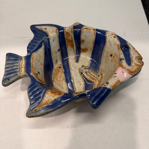 May include: A ceramic bowl shaped like a fish, featuring a cream-colored base with vertical blue stripes and brown speckled accents. The bowl has a defined tail, fins, and a small pink detail near the head, with a glossy finish throughout.