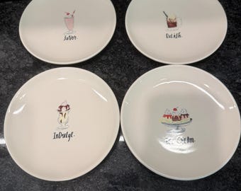 May include: A set of four round white ceramic plates, each featuring a unique dessert illustration and text. The designs include a pink milkshake labelled Savor, a brown drink labelled Delish, a sundae labelled Indulge, and a banana split labelled Ice Cream.