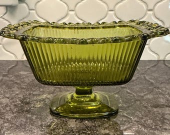 May include: A vintage, olive-green glass bowl with a square shape and a pedestal base. The bowl features vertical ribbed detailing on the sides and a beaded rim. The bowl is approximately 8 inches wide and 6 inches tall.