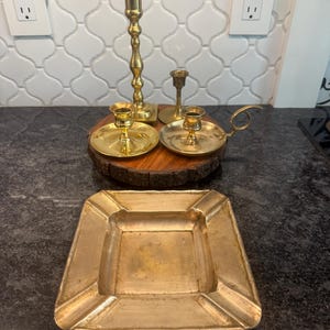 May include: A collection of vintage brass candlesticks and a square ashtray. The candlesticks are in various sizes and styles, some with ornate detailing. The ashtray has a square shape with slightly raised edges, showing signs of age.