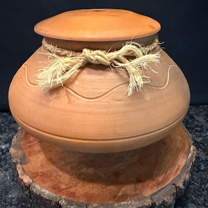 May include: A round terracotta pot with a matching lid, featuring a smooth surface with a subtle wavy line engraving around the center. A light-colored, fibrous rope is tied in a decorative knot around the neck of the pot, securing the lid in place.