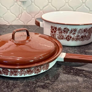 May include: A set of vintage-style enamelware cookware in white and brown. The set includes a frying pan with a lid, a casserole dish, and a small mug. The cookware features a floral pattern around the edges and on the casserole dish.