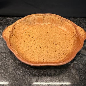 May include: A round ceramic pie dish with a scalloped edge and two integrated side handles. The dish features a speckled tan and brown glaze with a darker, mottled finish on the interior base and sides, creating a textured appearance.