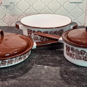 May include: A set of three vintage enamelware pots and pans. The cookware features a white base with a brown floral pattern and brown lids and handles. The set includes a large pot, a saucepan, and a covered oval dish.