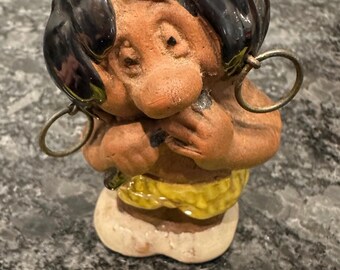 May include: A ceramic figurine of a person with black hair and large hoop earrings. The figure is brown with a yellow and white base. The figurine is approximately 5 inches tall.
