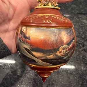 May include: A Christmas ornament with a winter scene. The ornament is a dark reddish-brown color with gold accents. The scene depicts deer in a snowy landscape with a house and trees. A small gold figurine of a horse-drawn carriage sits on top.