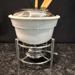 May include: A white ceramic fondue pot with a chrome metal stand and burner base. The set includes a chrome splash guard lid and four fondue forks with light-colored wooden handles. The pot sits securely within the circular metal frame structure.