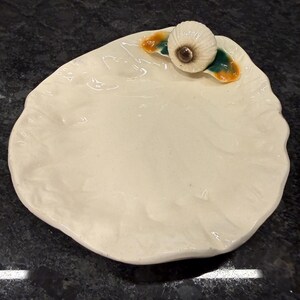 May include: Off-white ceramic oyster-shaped dish with a decorative seashell accent. The seashell is white with a brown center, and the dish has green and orange accents. The dish is on a dark surface.