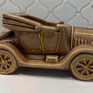 May include: A glossy tan ceramic planter shaped like a vintage convertible car. The vehicle features a folded roof, yellow-painted wheel rims, and yellow trim around the door frame. The open top of the car serves as a hollow container for plants.