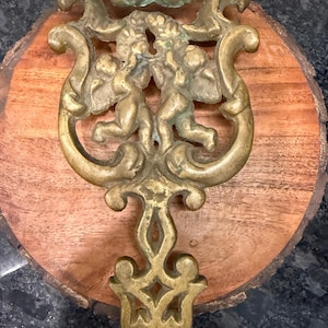May include: An antique brass wall sconce featuring two cherubs, ornate scrollwork, and a decorative base. The sconce is mounted on a round, natural wood slice. The brass has a weathered, aged appearance.