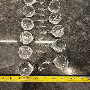 May include: Ten clear crystal prisms with faceted surfaces, hanging from a silver chain. The prisms are arranged in a vertical line, with the largest prism at the top and the smallest prism at the bottom. A yellow measuring tape is visible at the bottom of the image.