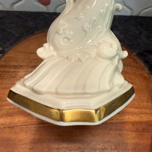 May include: Ivory-colored decorative porcelain figurine with gold trim. The figurine has an ornate, flowing design with detailed relief patterns. The base is a geometric shape with a gold border. The figurine is placed on a wooden surface.