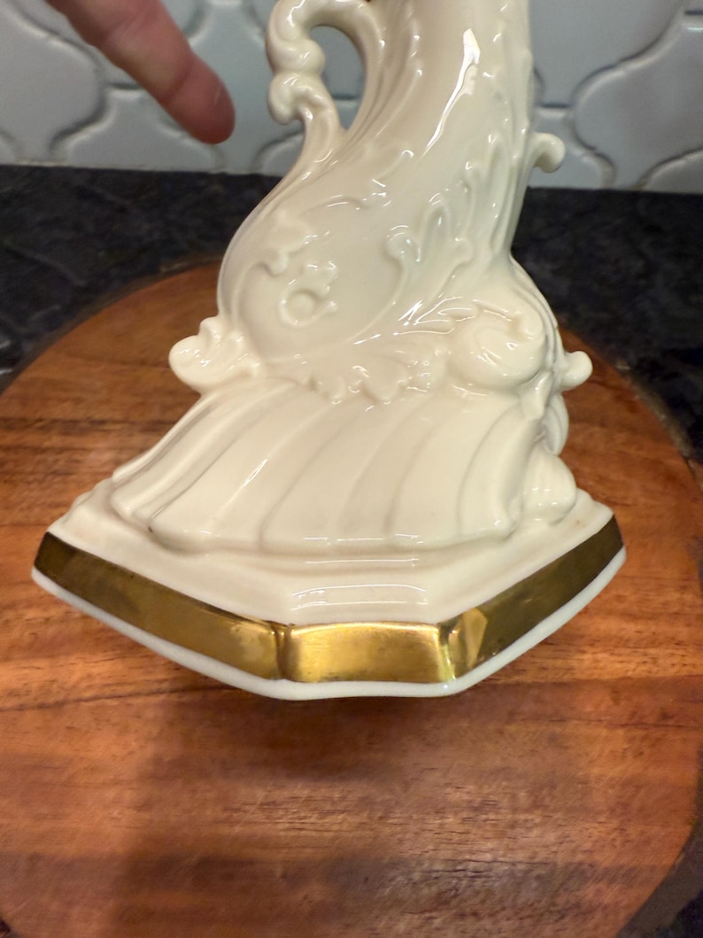 May include: Ivory-colored decorative porcelain figurine with gold trim. The figurine has an ornate, flowing design with detailed relief patterns. The base is a geometric shape with a gold border. The figurine is placed on a wooden surface.