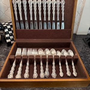 May include: A wooden silverware chest with a set of ornate silver-colored flatware. The set includes knives, forks, and spoons, all neatly arranged in a brown velvet-lined interior. The chest is open, showcasing the complete service.