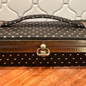 May include: A vintage black handbag with a handle and gold-colored hardware. The bag is covered in small, evenly spaced yellow polka dots. The edges are trimmed with gold-colored metal. The bag is rectangular and sits on a wooden surface.