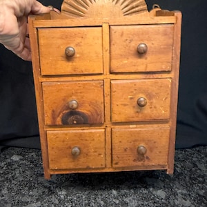 May include: A wooden wall-mounted spice cabinet featuring six small drawers with round knobs. The top of the cabinet has a decorative semi-circular sunburst carving. The wood has a natural light brown finish with visible grain and knots.