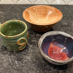 May include: Three handmade ceramic bowls and a mug. The mug is green with a brown handle and decorative accents. One bowl is orange, and the other is blue and red. The pottery has a rustic, artisanal appearance.