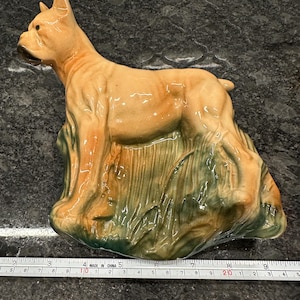 May include: A ceramic figurine of a boxer dog standing in a bed of green and brown grass. The dog is light brown with a white chest and is standing with its head turned to the side. The figurine is made in China.