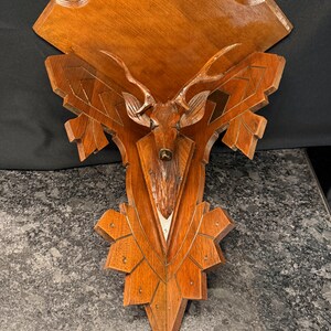 May include: A wall-mounted wooden plaque featuring a carved deer head with antlers at the center. The plaque has a polished brown finish with geometric, angular edges and decorative carved lines radiating outward from the central deer figure.