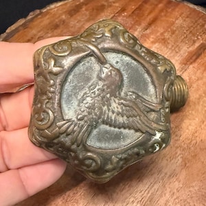 May include: A tarnished brass faucet handle featuring a six-pointed star shape with ornate scrollwork along the edges. The center of the handle displays a raised relief image of a bird in flight, and a threaded metal stem extends from the back.