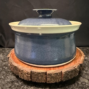 May include: A round ceramic casserole dish with a deep blue speckled glaze and a matching lid. The pot features a wide, flared rim and a central knob handle on the lid. The interior and the underside of the rim are finished in a smooth, off-white glaze.