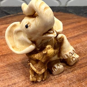 May include: Ivory-colored elephant figurine with trunk raised and curled. The elephant is detailed with visible wrinkles and a textured surface. The figurine is posed as if it is holding onto a tree trunk. The elephant is on a wooden surface.