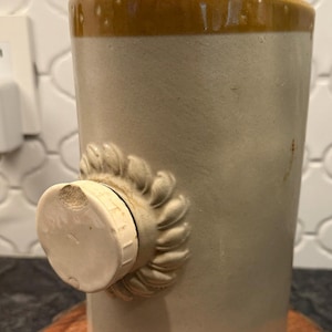May include: A vintage stoneware hot water bottle with a tan and cream color scheme. The bottle has a decorative sunflower-shaped stopper and a matching lid. The bottle is cylindrical and sits on a wooden surface.