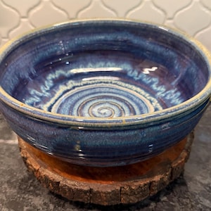 May include: A round ceramic bowl featuring a deep blue glaze with lighter blue and white swirling patterns on the interior. The bowl has a thick, rounded rim and small handles on opposite sides, showcasing a glossy finish and a spiral design.